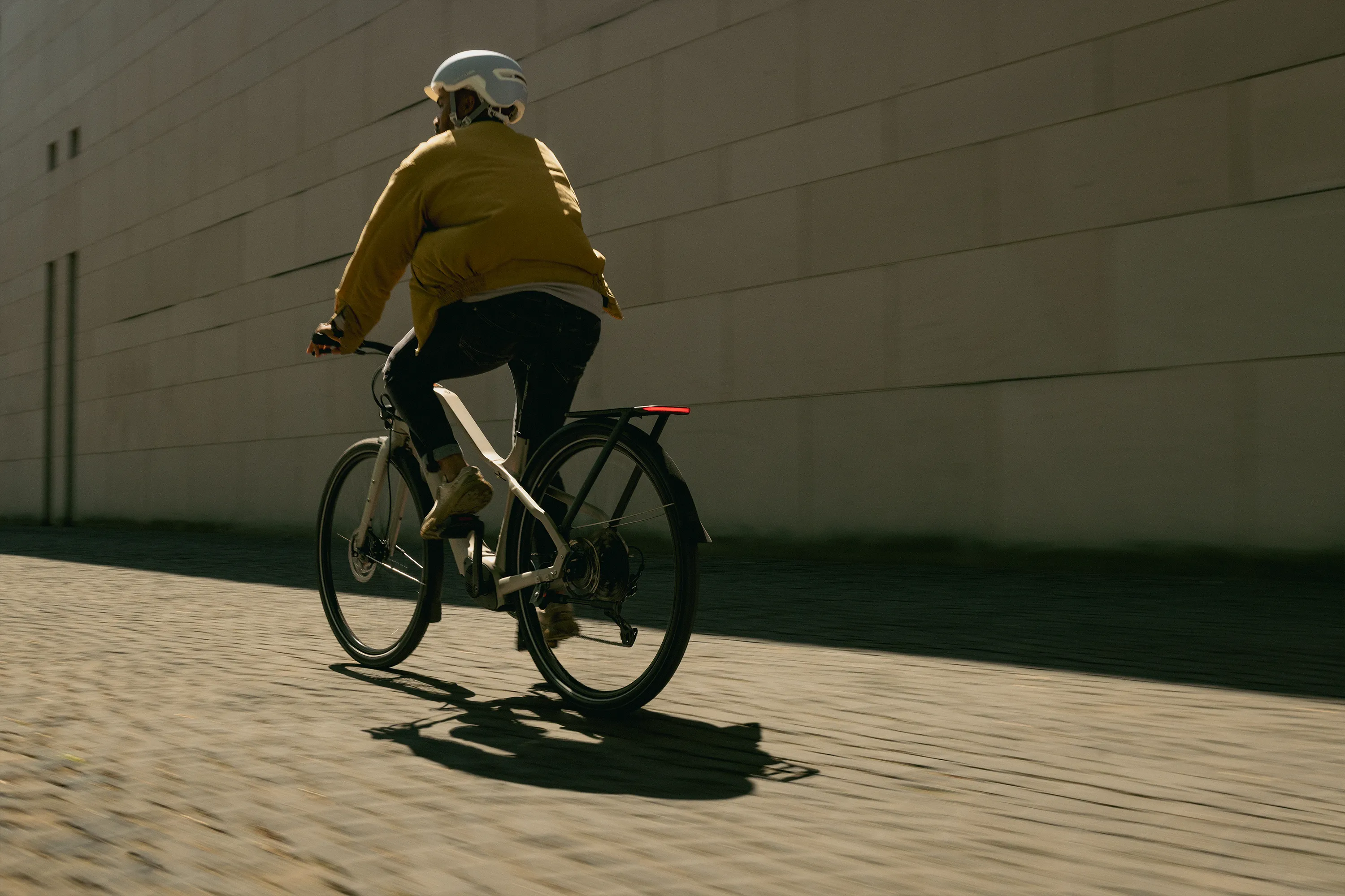 Salarié casqué qui se rend au travail sur son vélo de fonction, de dos, le long d'un mur au soleil