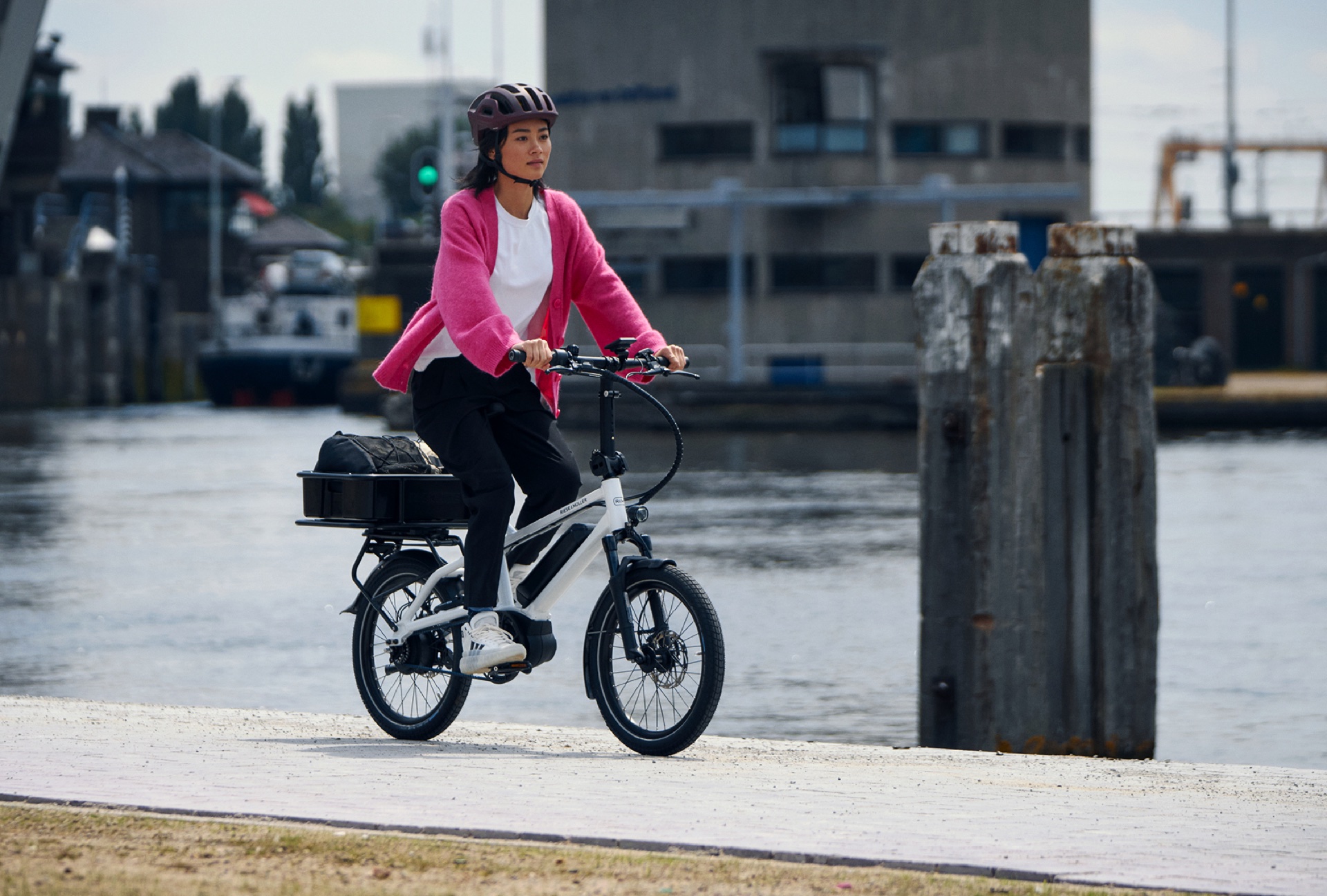 Salariée casquée qui longe un quai sur un vélo électrique compact, un sac dans le panier arrière.