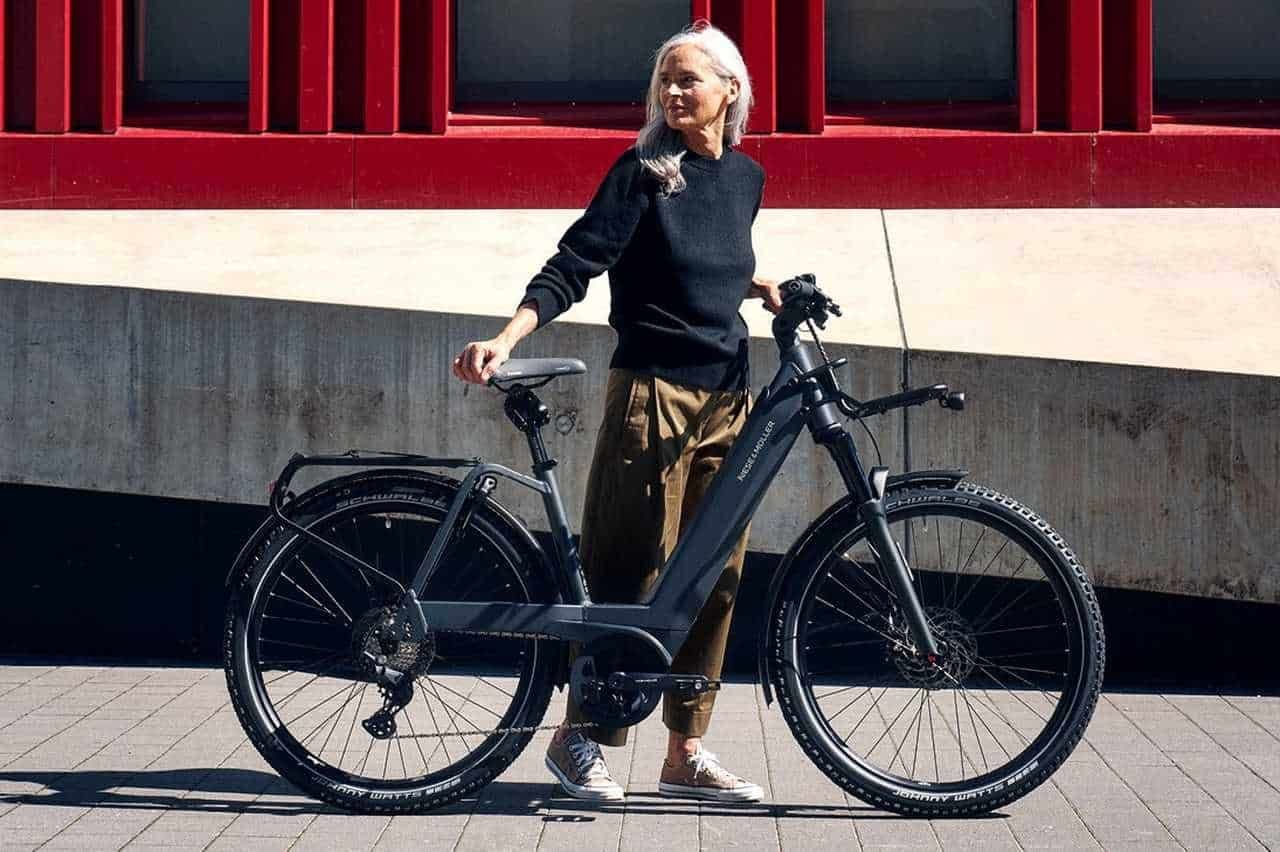 Woman with gray hair in a black sweater and olive pants stands with a black electric bicycle.