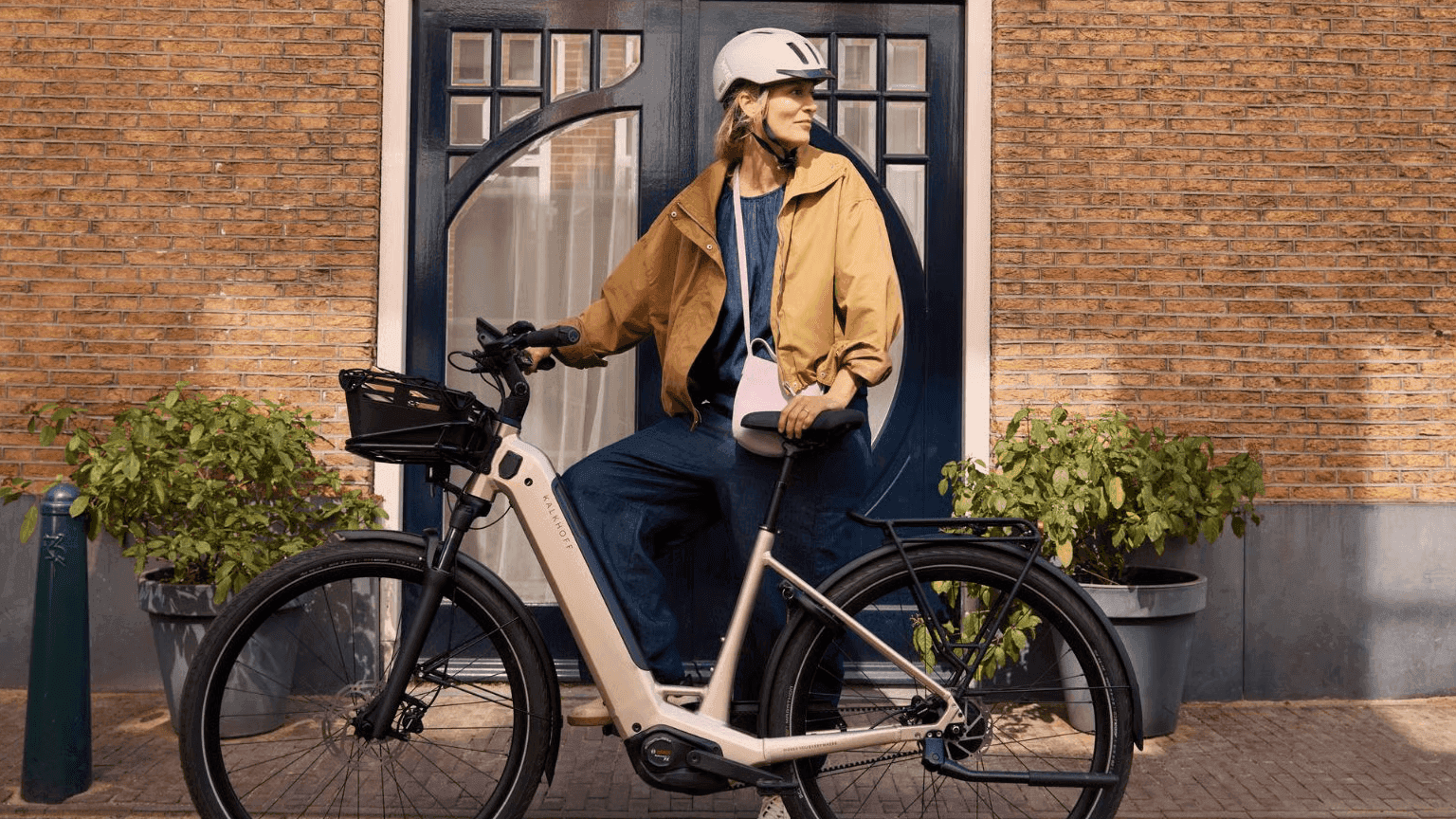 A woman in a helmet and jacket stands with an electric bicycle in front of a brick building.